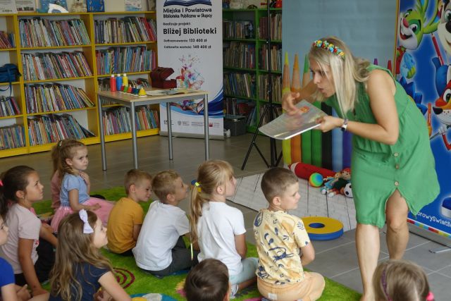 dzieci siedzą na dywanie, przed nimi stoi kobieta i zdmuchuje pyłki z książki, w lewym rogu duży plakat z nazwą projektu i logo biblioteki na rozłożonej książce herb Słupcy na niebieskim tle żółty krzyż oraz herb powiatu słupeckiego oraz herb powiatu słupeckiego, od góry na czerwonym tle biały orzeł do połowy, od dołu na niebieskim tle wokoło małe żółte krzyże