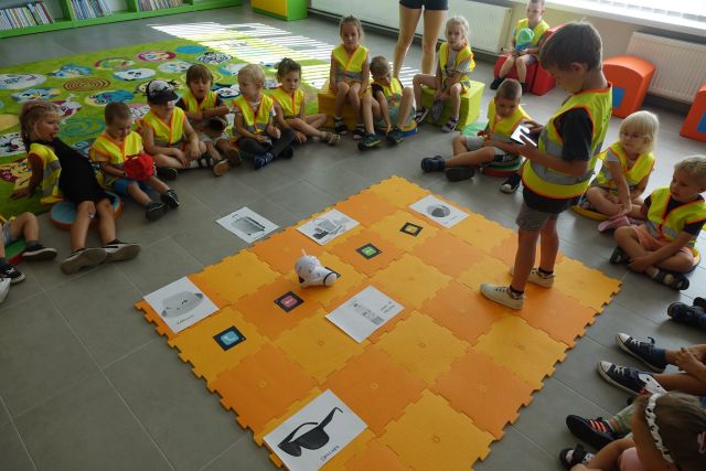 dzieci w zielonych kamizelkach siedzą wokół rozłożonej na podłodze pomarańczowej macie, po której jeździ robot, którym steruje z tabletu chłopiec
