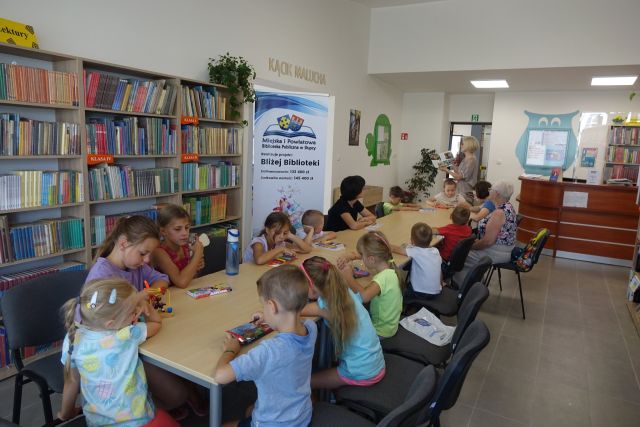 przy brązowych stołach siedzą dzieci i słuchają czytanej przez kobietę książki, z lewej strony stoi duży plakat z nazwą projektu i logo biblioteki na rozłożonej książce herb Słupcy na niebieskim tle żółty krzyż oraz herb powiatu słupeckiego oraz herb powiatu słupeckiego, od góry na czerwonym tle biały orzeł do połowy, od dołu na niebieskim tle wokoło małe żółte krzyże