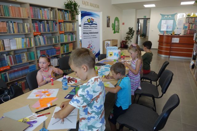 przy brązowych stołach złączonych siedzą dzieci i malują na kartkach, za nimi brązowe regały z książkami i duży plakat z nazwą projektu i logo biblioteki na rozłożonej książce herb Słupcy na niebieskim tle żółty krzyż oraz herb powiatu słupeckiego oraz herb powiatu słupeckiego, od góry na czerwonym tle biały orzeł do połowy, od dołu na niebieskim tle wokoło małe żółte krzyże