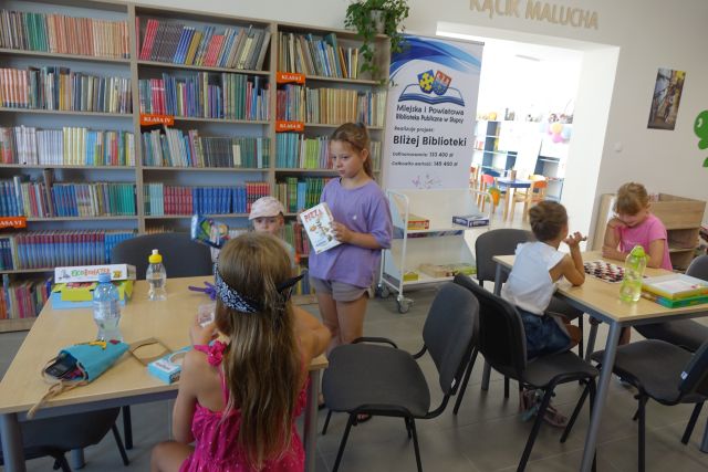 przy brązowych stołach dzieci grają w gry planszowe, za nimi stoi duży plakat z nazwą projektu i logo biblioteki na rozłożonej książce herb Słupcy na niebieskim tle żółty krzyż oraz herb powiatu słupeckiego oraz herb powiatu słupeckiego, od góry na czerwonym tle biały orzeł do połowy, od dołu na niebieskim tle wokoło małe żółte krzyże