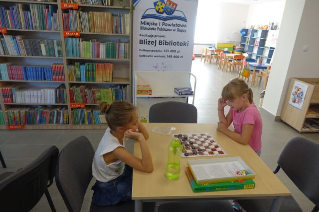 przy brązowym stole siedzi chłopiec i dziewczynka i grają w gry planszowe, za nimi regały z książkami i duży plakat z nazwą projektu i logo biblioteki na rozłożonej książce herb Słupcy na niebieskim tle żółty krzyż oraz herb powiatu słupeckiego oraz herb powiatu słupeckiego, od góry na czerwonym tle biały orzeł do połowy, od dołu na niebieskim tle wokoło małe żółte krzyże
