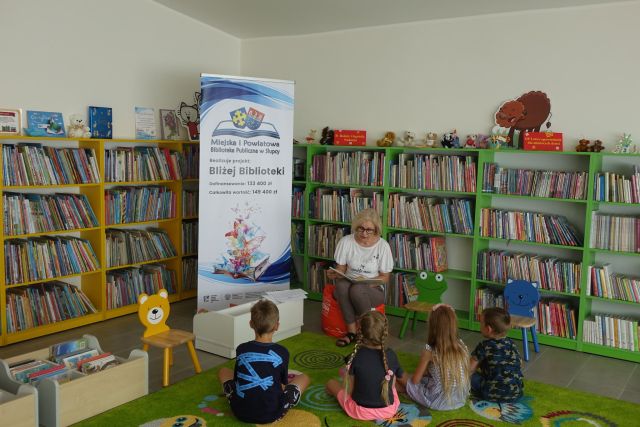 na zielonym dywanie siedzą dzieci, przed nimi na krzesełku siedzi kobieta i czyta książkę, za nimi zielone i żółte regały z książkami oraz duży plakat z nazwą projektu i logo biblioteki na rozłożonej książce herb Słupcy na niebieskim tle żółty krzyż oraz herb powiatu słupeckiego oraz herb powiatu słupeckiego, od góry na czerwonym tle biały orzeł do połowy, od dołu na niebieskim tle wokoło małe żółte krzyże