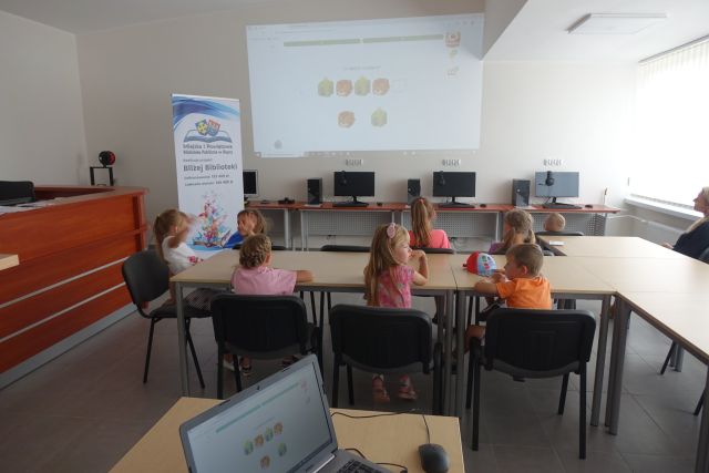 przy brązowych stołach siedzą dzieci, przed nimi na ekranie wyświetlają się zagadki, z lewej strony stoi duży plakat z nazwą projektu i logo biblioteki na rozłożonej książce herb Słupcy na niebieskim tle żółty krzyż oraz herb powiatu słupeckiego oraz herb powiatu słupeckiego, od góry na czerwonym tle biały orzeł do połowy, od dołu na niebieskim tle wokoło małe żółte krzyże