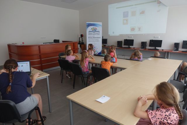 pzry stołąch siedzą dzieci, na ekranie przed nimi wyświetlają się zagadki, z lewej strony stoi duży plakat z nazwą projektu i logo biblioteki na rozłożonej książce herb Słupcy na niebieskim tle żółty krzyż oraz herb powiatu słupeckiego oraz herb powiatu słupeckiego, od góry na czerwonym tle biały orzeł do połowy, od dołu na niebieskim tle wokoło małe żółte krzyże