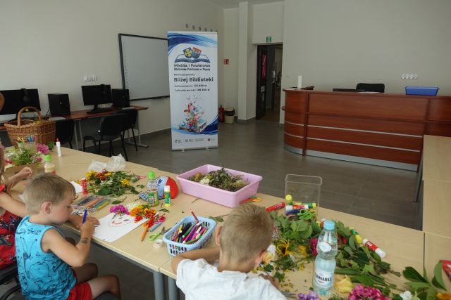 przy brązowych stołach siedzą dzieci i wykonują rysunki i wyklejanki z kwiatów