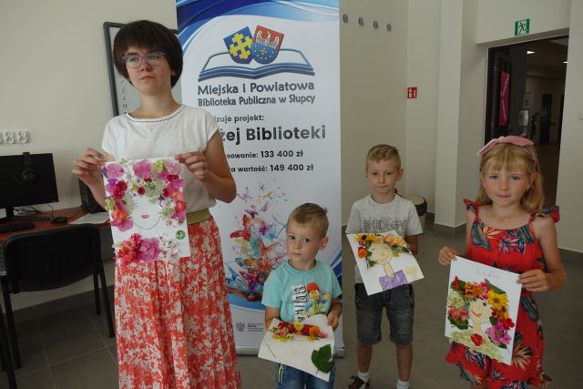 dzieci stoją przed dużym plakatem z nazwą projektu i logo biblioteki na rozłożonej książce herb Słupcy na niebieskim tle żółty krzyż oraz herb powiatu słupeckiego oraz herb powiatu słupeckiego, od góry na czerwonym tle biały orzeł do połowy, od dołu na niebieskim tle wokoło małe żółte krzyże, dzieci w rękach trzymaja wykonane przez siebie plakaty z twarzami z włosami wyklejonymi z kwiatów