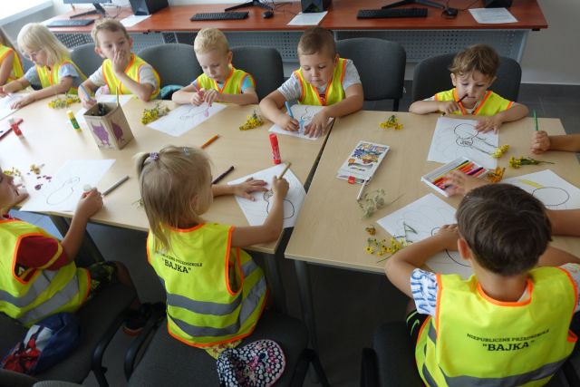przy brązowych stołach siedzą dzieci w zielonych kamizelkach i rysują na kartkach