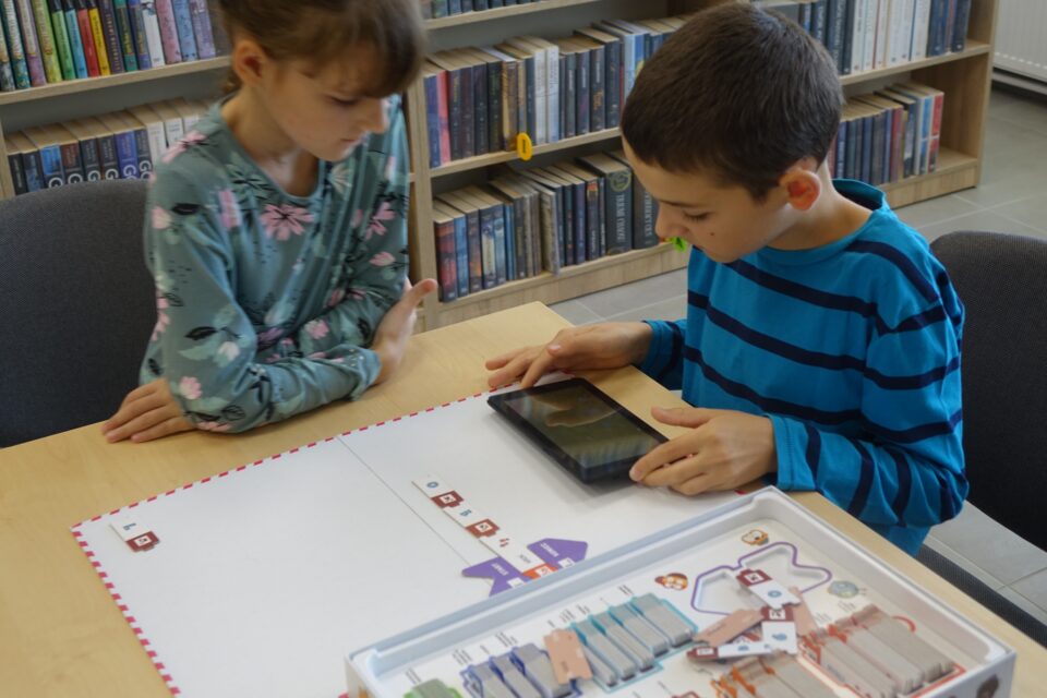 dwóch chłopców siedzi przy stole, na stole leży gra, tablet i kartka papieru