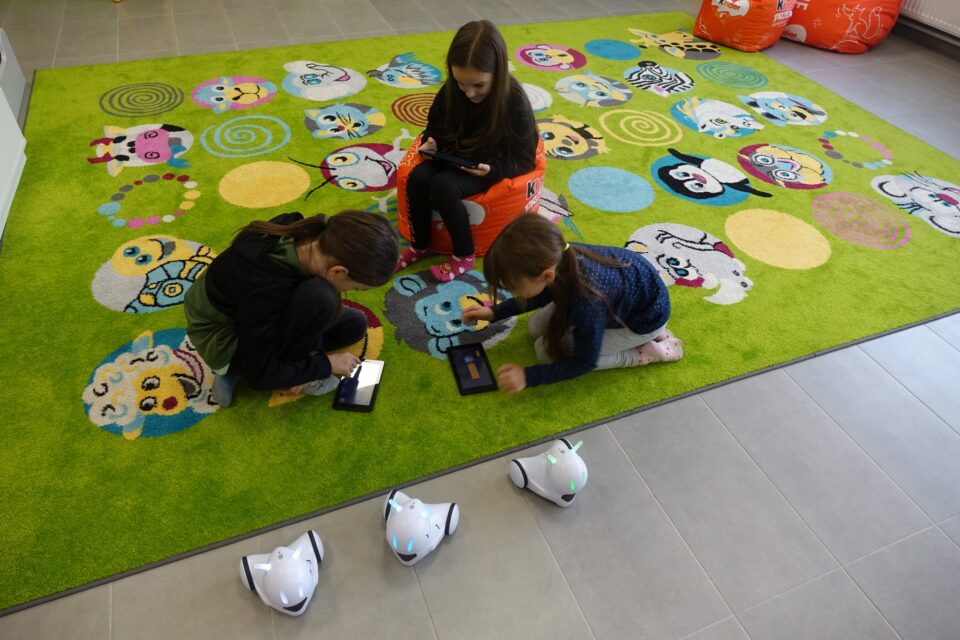 trójka dzieci siedzą na zielonym dywanie i sterują poprzez tablety małymi białymi robotami