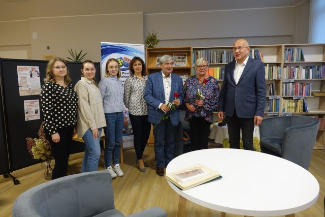 uczestnicy spotkania stoją, kobieta w niebieskiej bluzce we wzory i mężczyzna w siwym garniturze trzymają czerwone róże