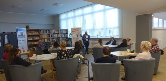 uczestnicy spotkania siedzą na siwych fotelach przy białych stolikach, mężczyzna w siwym garniturze stoi, kobieta w bluzce niebieskiej we wzory siedzi, w tle pomieszczenie biblioteczne