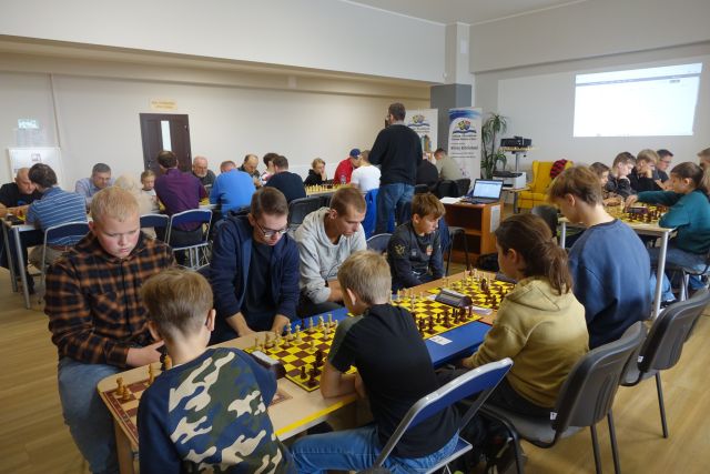 uczestnicy turnieju siedzą przy stołach i grają w szachy, figurki białe i czarne na brązowo-żółtych planszach