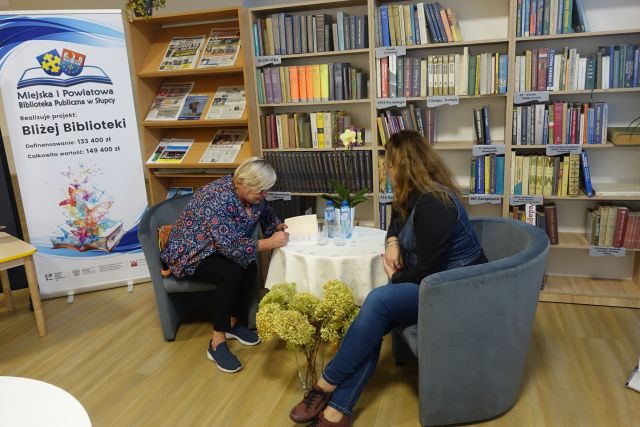 dwie kobie siedzą przy małym stoliku z kremowym obrusem, w tle regały z książkami