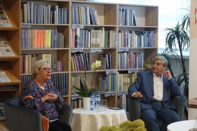 kobieta w bluzce niebieskiej we wzory i mężczyzna w siwym garniturze i białej koszuli siedzą przy stoliku z kremowym obrusem, w tle regały z książkami