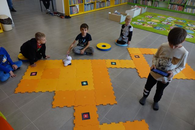 dzieci siedzą na podłodze, na której rozłożone są pomarańczowe duże piankowe puzzle, przed nimi mały biały robot, chłopiec stoi i trzyma w rękach tablet