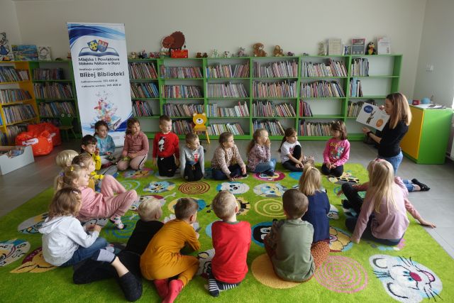 dzieci i prowadząca siedzą na zielonym dywanie, za nimi zielone regały z książkami