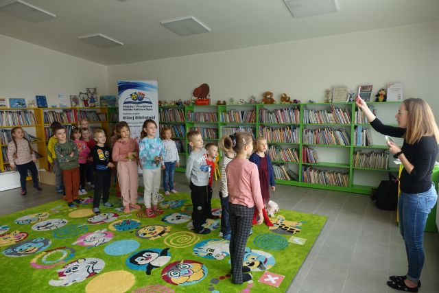 dzieci stoją na zielonym dywanie, za nimi zielone regały z książkami