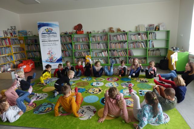 dzieci siedzą na zielonym dywanie, w tle zielone regały z książkami