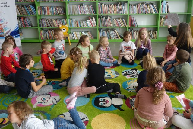 dzieci i prowadząca z książką siedzą na zielonym dywanie, w tle zielone regały z książkami