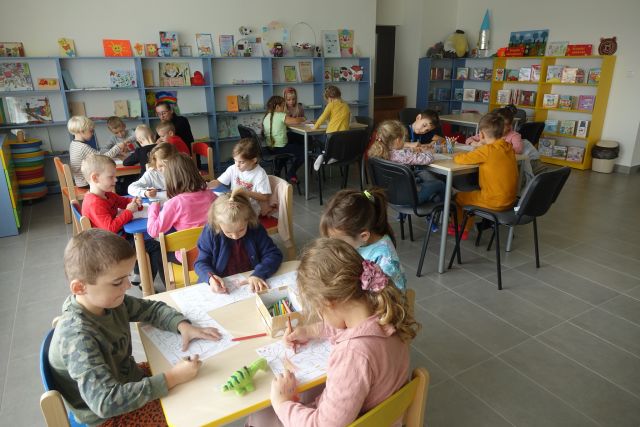 dzieci siedzą przy stolikach, w tle niebieskie regały