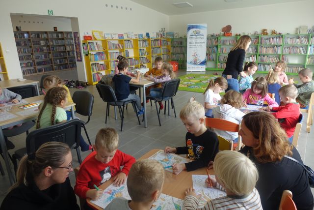 dzieci siedzą przy stolikach, w tle żółte i zielone regały z książkami