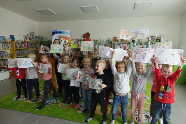 dzieci stoją na zielonym dywanie i tzrymają w rękach rysunki, w tle zielone regały z książkami