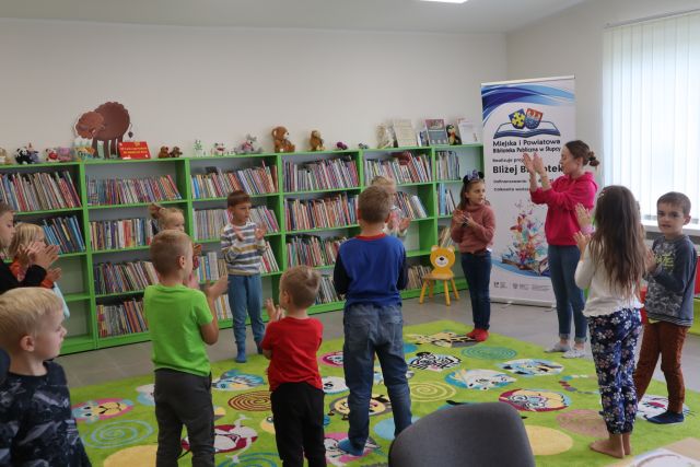 dzieci kolorowo ubrane stoją na zielonym dywanie, kobieta w czerwonej bluzce pokazuje im ręce, za nimi zielone regały z książkami