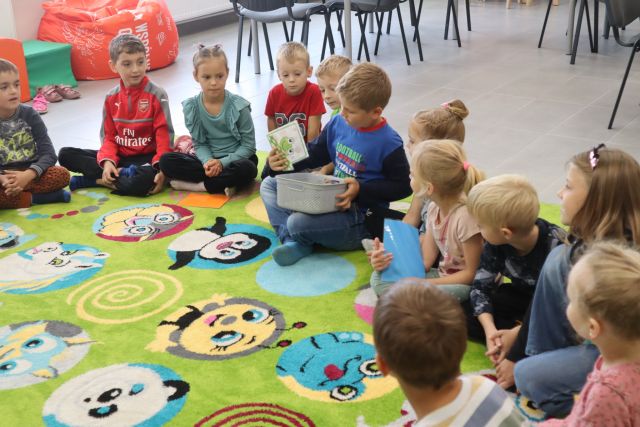 Dzieci siedzą na zielonym dywanie, chłopiec w niebieskiej bluzce trzyma na kolanach pudełko z kartkami