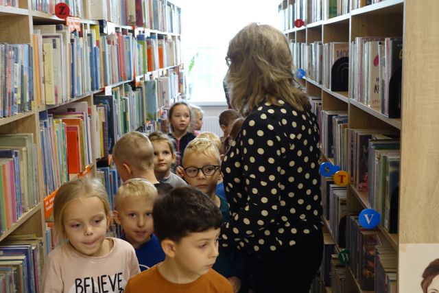 dzieci i dorosła kobieta w czarnej bluzce w kropki stoją pomiędzy regałami z książkami