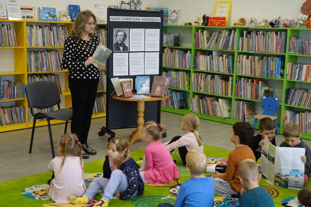 dzieci siedzą na zielonym dywanie, kobieta w czarnej bluzce w kropki stoi, w tle zielone i żółte regały z książkami