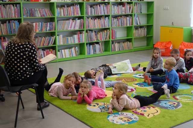 dzieci leżą na zielonym dywanie, przed nimi siedzi na krześle kobieta w czarnej bluzce w kropki i czarnych spodniach, w tle zielone regały z książkami