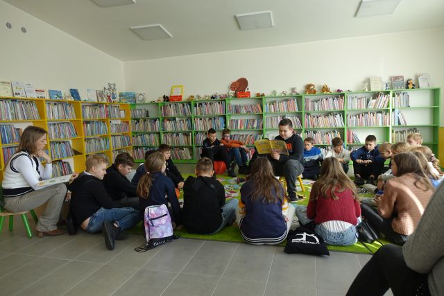 młodzież siedzi na zielonym dywanie, chłopiec w środku siedzi na krześle i czyta książkę, w tle regały z książkami