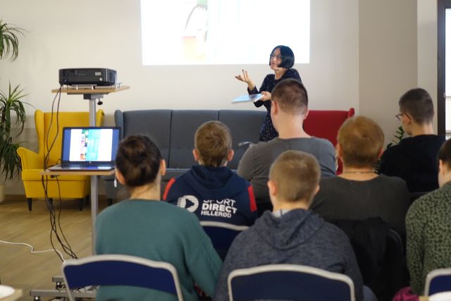 młodzież siedzi tyłem do aparatu, przed nimi stoi kobieta, w tle biały ekran