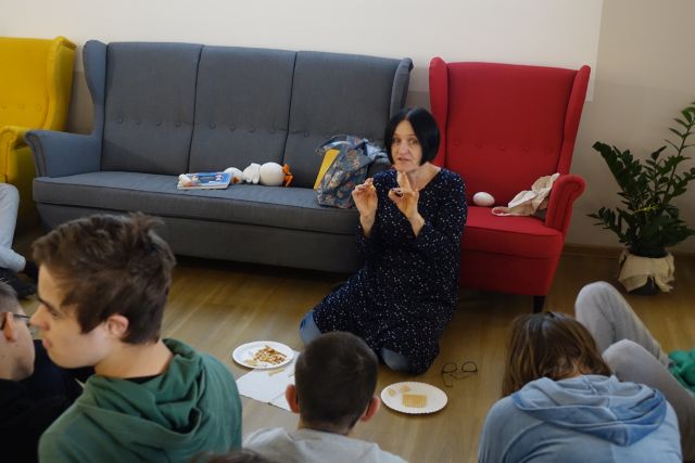 kobieta ubrana w ciemne kolory bluzkę i spodnie siedzi na podłodze, za nią czerwony fotel i siwa sofa