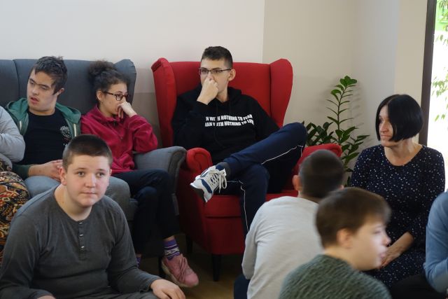 na podłodze siedzi młodzież, na czerwonym fotelu siedzi chłopiec w okularach i czarnej bluzie, na siwej sofie siedzą chłopcy w czerwonej bluzce i siwej bluzce