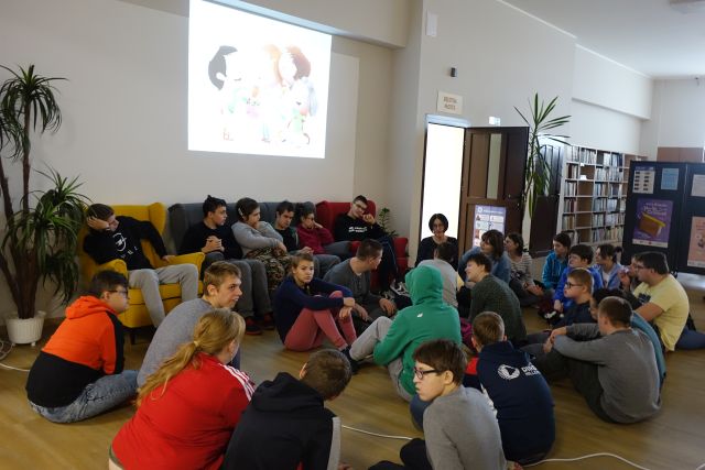 młodzież siedzi na podłodze, między nimi siedzi kobie ubrana w ciemne kolory bluzkę i spodnie, w tle na sofie siedzą chłopcy