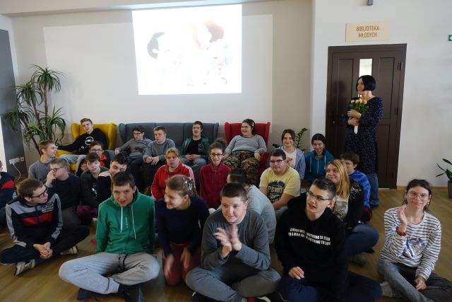 młodzież siedzi na podłodze, z prawej strony za nimi stoi kobieta z żółtymi różami