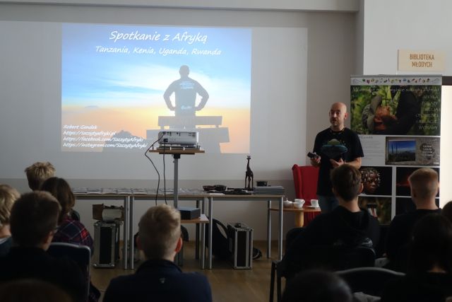 młodzież siedzi na krzesłąch, przed nimi stoi mężczyzna w czarnej bluzce, na ekranie wyświetlane są slajdy ze zdjęciami z Afryki
