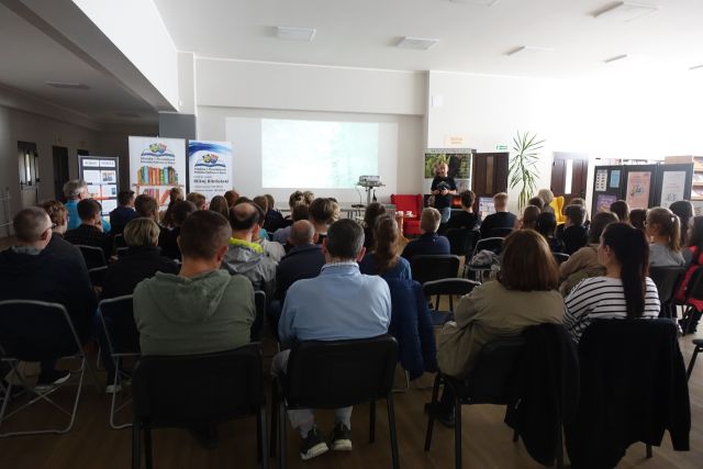 uczestnicy spotkania siedzą na krzesłąch, przed nimi mężczyzna w czarnej bluzce i biały ekran