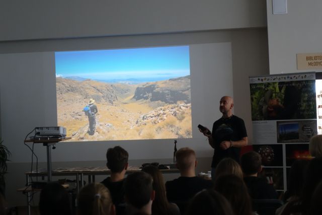 mężczyzna w czarnej bluzce stoi przed ekranem ze zdjęciami z Afryki, przed nim na krzesłąch siedzą uczestnicy spotkania