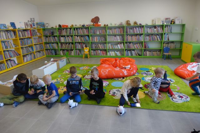 na zielonym dywanie siedzą dzieci i sterują małymi białymi robotami, w tle zielone regały z książkami