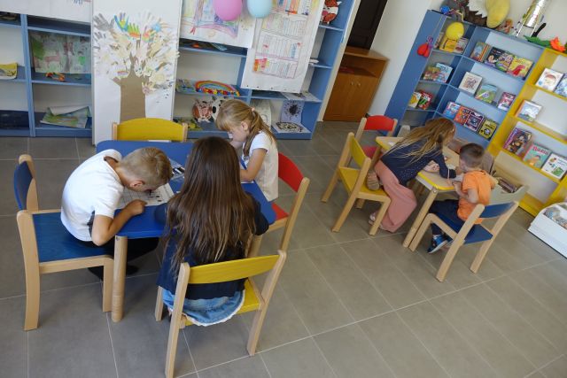 przy małych kolorowych stołąch siedzą dzieci, na stołach leżą kartki, za stołami żółte i niebieskie regały