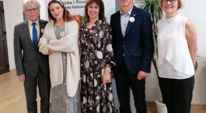 mężczyzna w siwym garniturze, kobieta w jasnej sukience i biały swetrze, kobieta w granatowej sukience w kwiaty, mężczyzna w granatowym garniturze, kobieta w białej bluzce i czarnych spodniach stoją