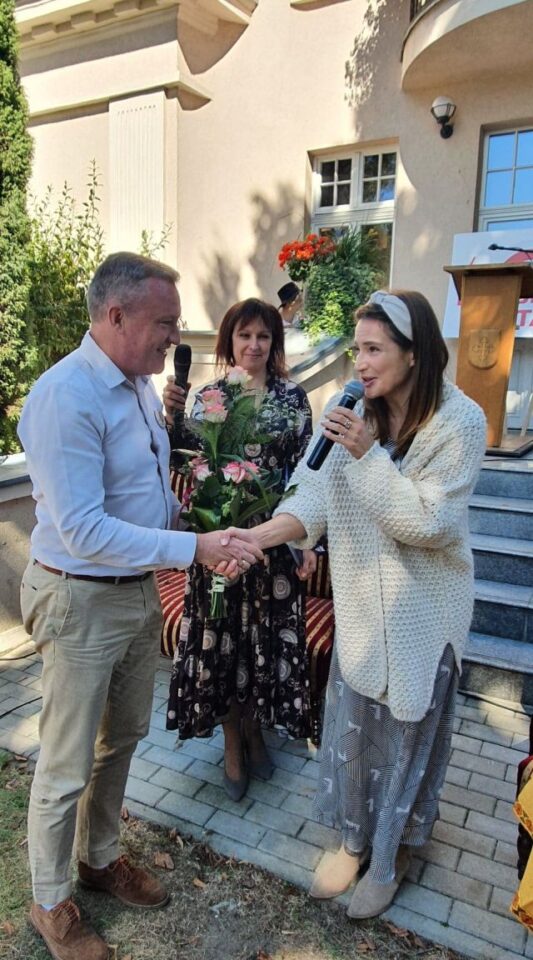 mężczyzna w białej koszuli i jasnych spodniach wręcza kobiecie w jasnej sukience i białym swetrze kwiaty, pomiędzy nimi z tyłu stoi kobieta w ciemnej sukience w kwiaty