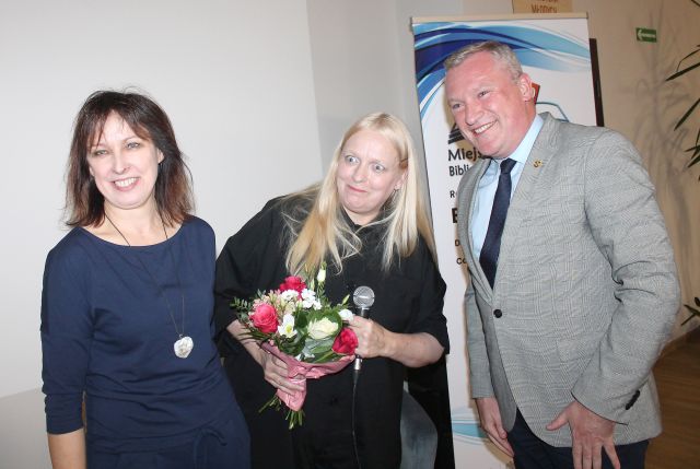 mężczyzna w jasnym garniturze, kobieta w czarnym kombinezonie z bukietem kolorowych kwiatów, kobieta w granatowym kombinezonie