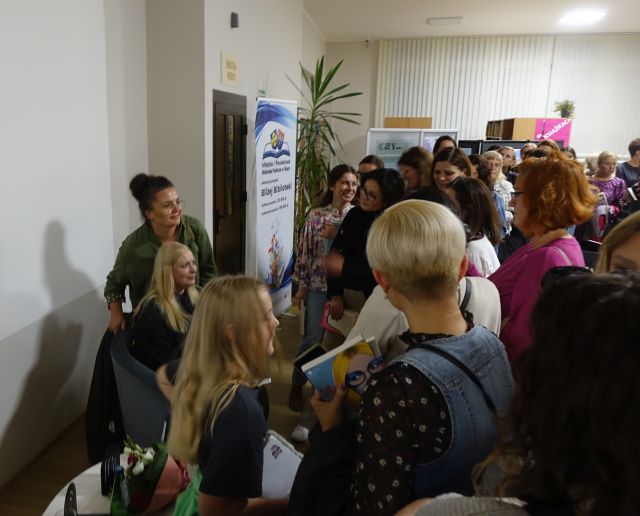 tłum ludzi stoi w kolejce po autograf, kobieta w czarnym kombinezonie siedzi i pozuje do zdjęcia