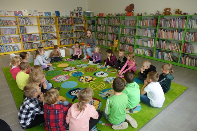 dzieci siedzą na zielonym dywanie w kole i kobieta z książką, za nimi zielone i żółte regały z książkami