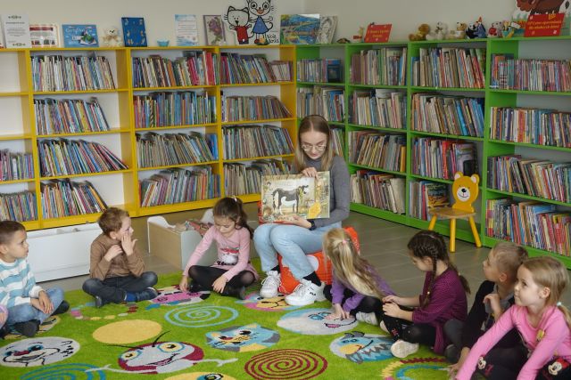 kobieta w siwej bluzce siedzi na czerwoje pufie i trzyma w rękach książkę, dzieci siedzą na zielonym dywanie, za nimi zielone i żółte regały z książkami