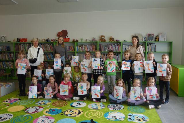 dzieci i dwie dorosłe osoby w siwej bluzce i różowej stoją na zielonym dywanie i trzymają w rękach rysunki na kartkach